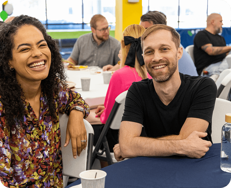 A woman and a man smiling at a table during a casual gathering, with other people sitting in the background.
