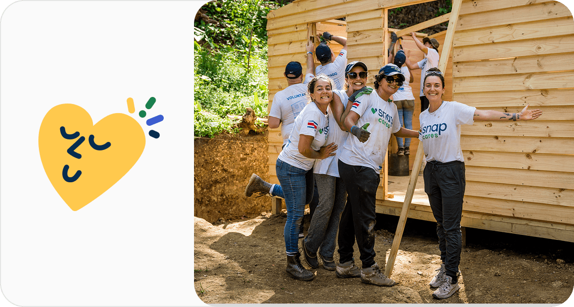 Volunteers in white T-shirts smiling while building a wooden structure outdoors; heart icon on the left.