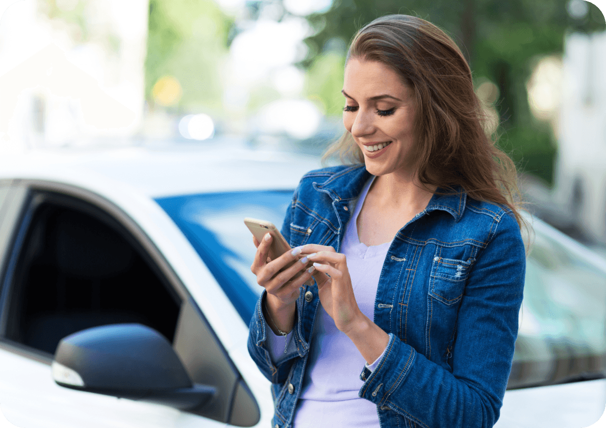 Woman in a denim jacket smiling while using a smartphone, standing beside a white car on a sunny day.