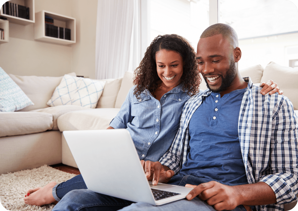 A smiling couple sits on the floor by a sofa, looking at a laptop together in a cozy living room.
