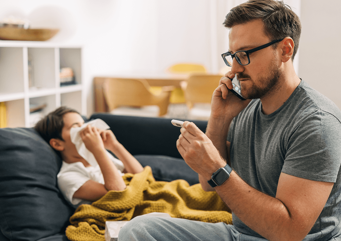 Man on phone checks a thermometer, seated beside a child with a tissue and yellow blanket on a sofa.