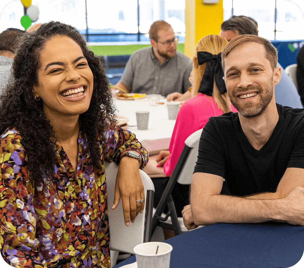 Two people smiling and sitting side by side at a table during a social event. Other attendees are visible in the background.