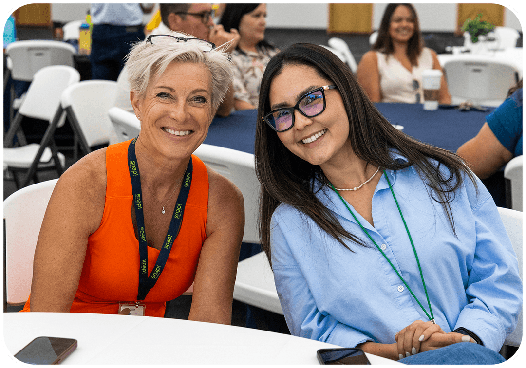 Two women smiling at a table during a casual event, surrounded by others seated at tables in the background.
