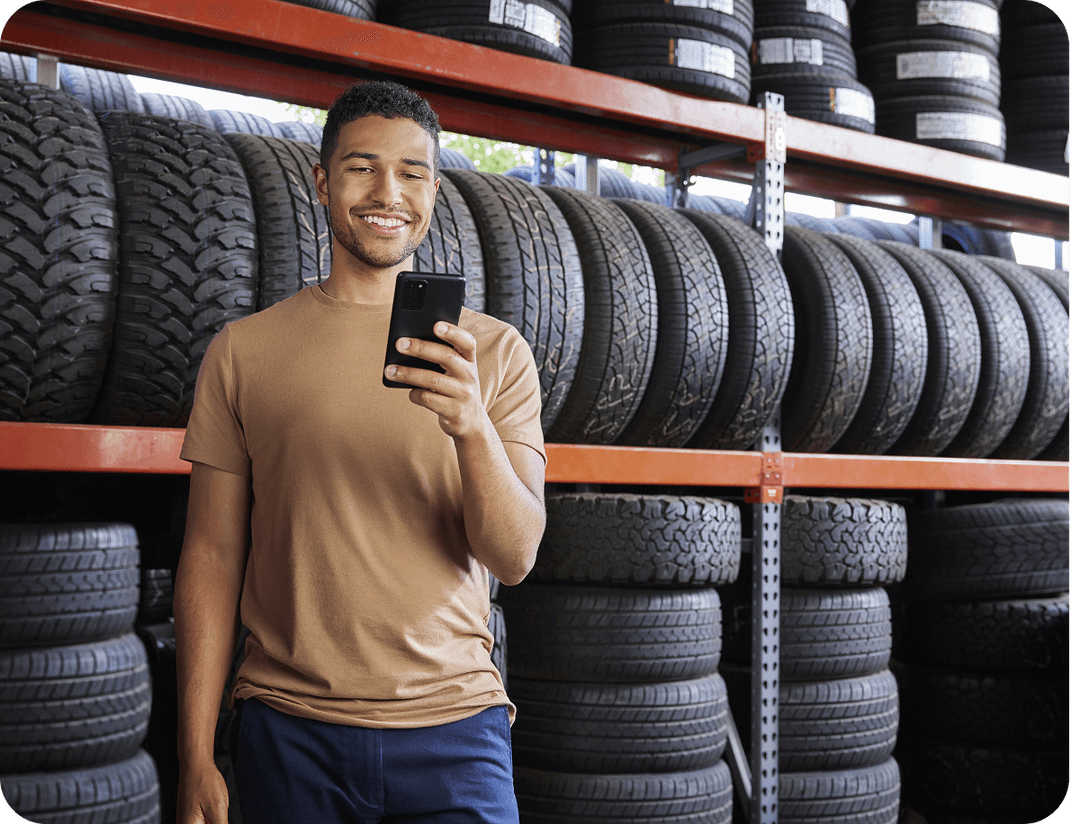 Man in a beige shirt smiles at his phone, standing in front of stacked tires on orange shelves in a store.