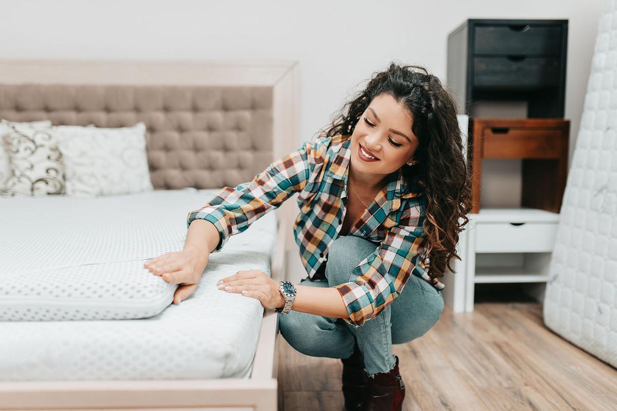 Woman kneels beside showroom bed, testing a mattress topper’s firmness with her hand while smiling in evaluation.