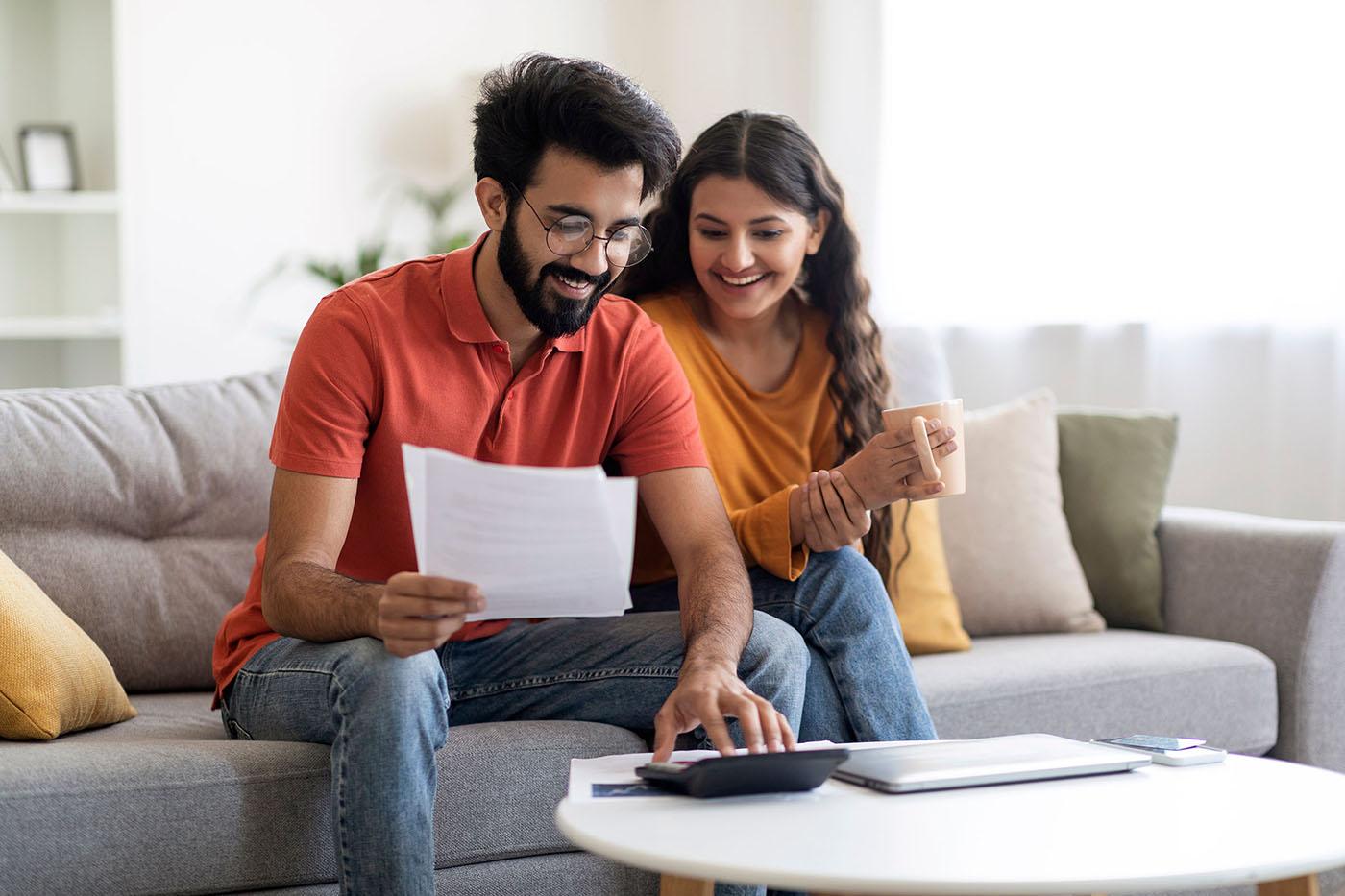 A smiling couple sits on a couch reviewing documents together; the man holds papers while using a calculator and the woman holds a coffee mug.