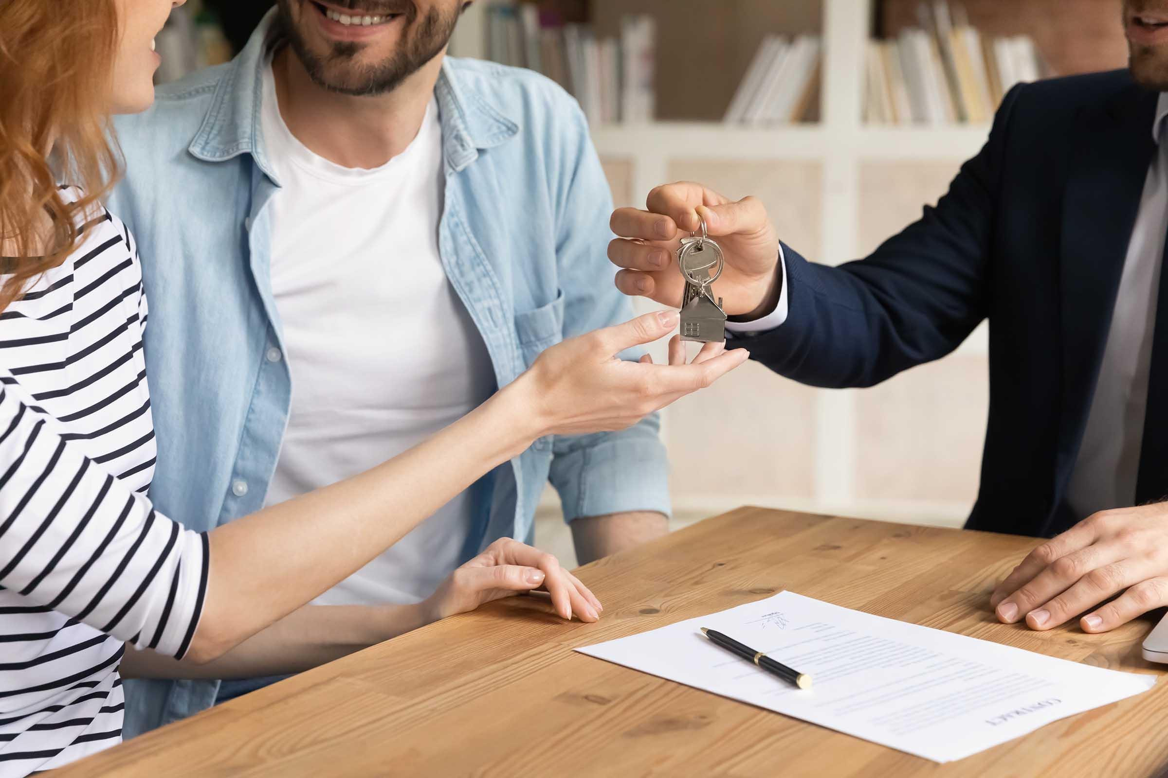 Real estate agent handing house keys to a smiling couple across a table with a signed contract and pen.
