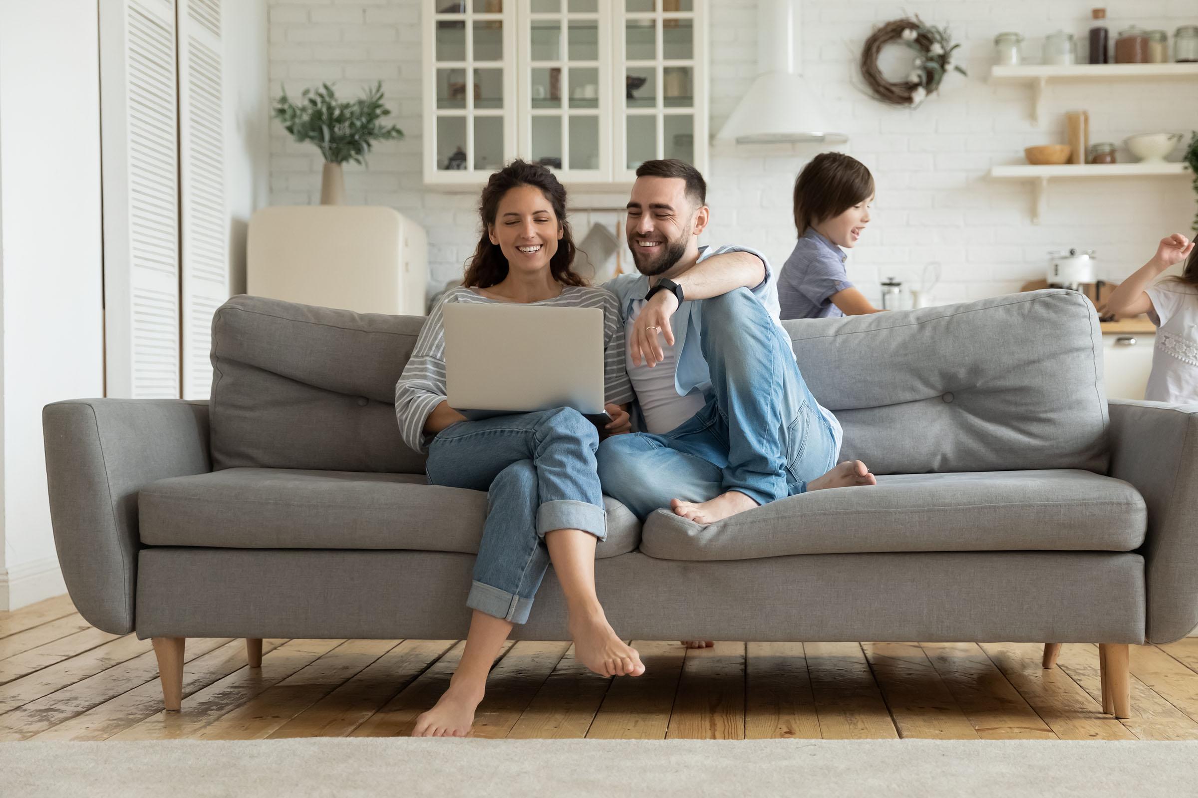 Smiling couple sitting on a gray sofa using a laptop, with two children playing in the background of a bright, modern living room.