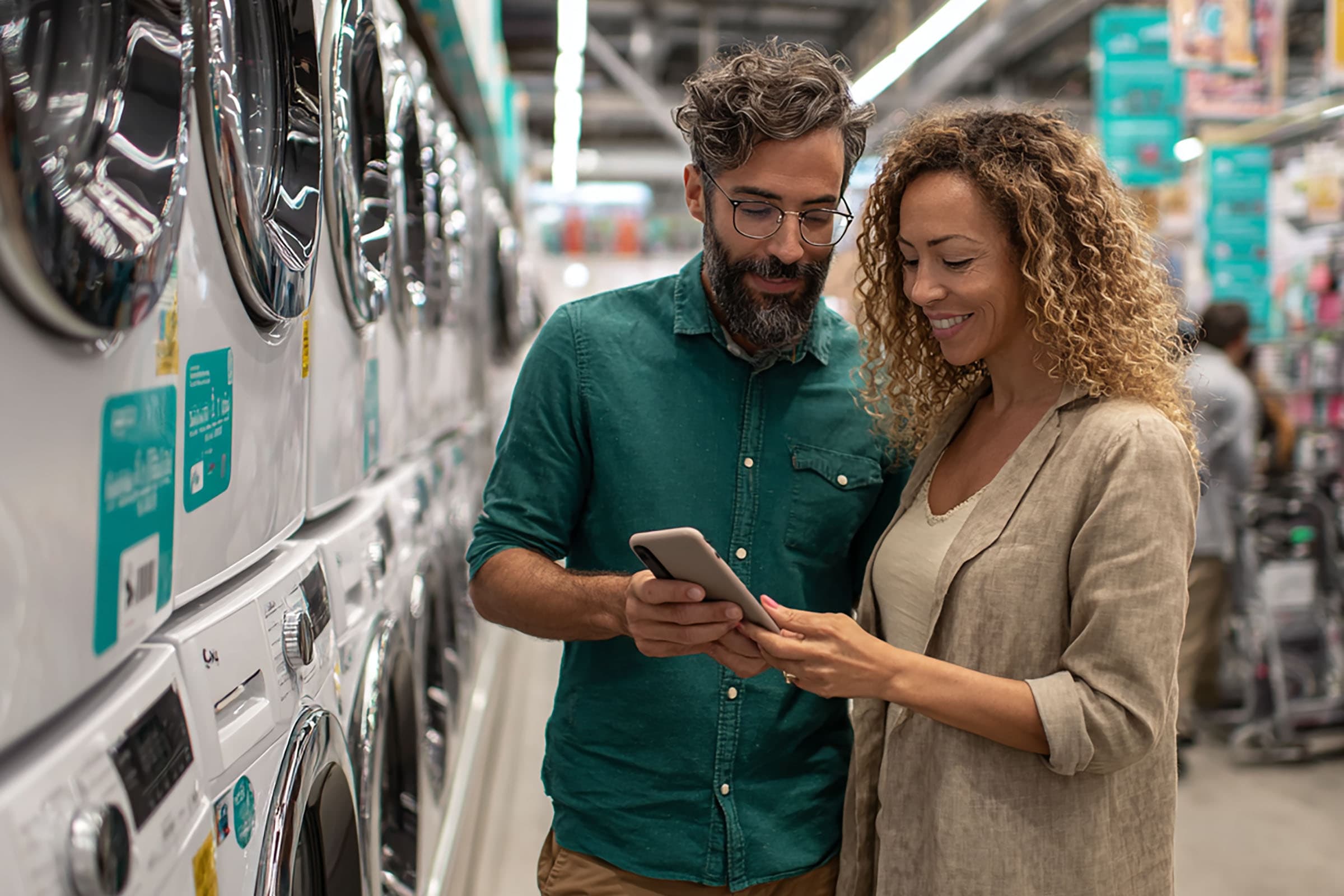 A man and woman smile while looking at a smartphone in an appliance store aisle lined with washing machines and dryers.