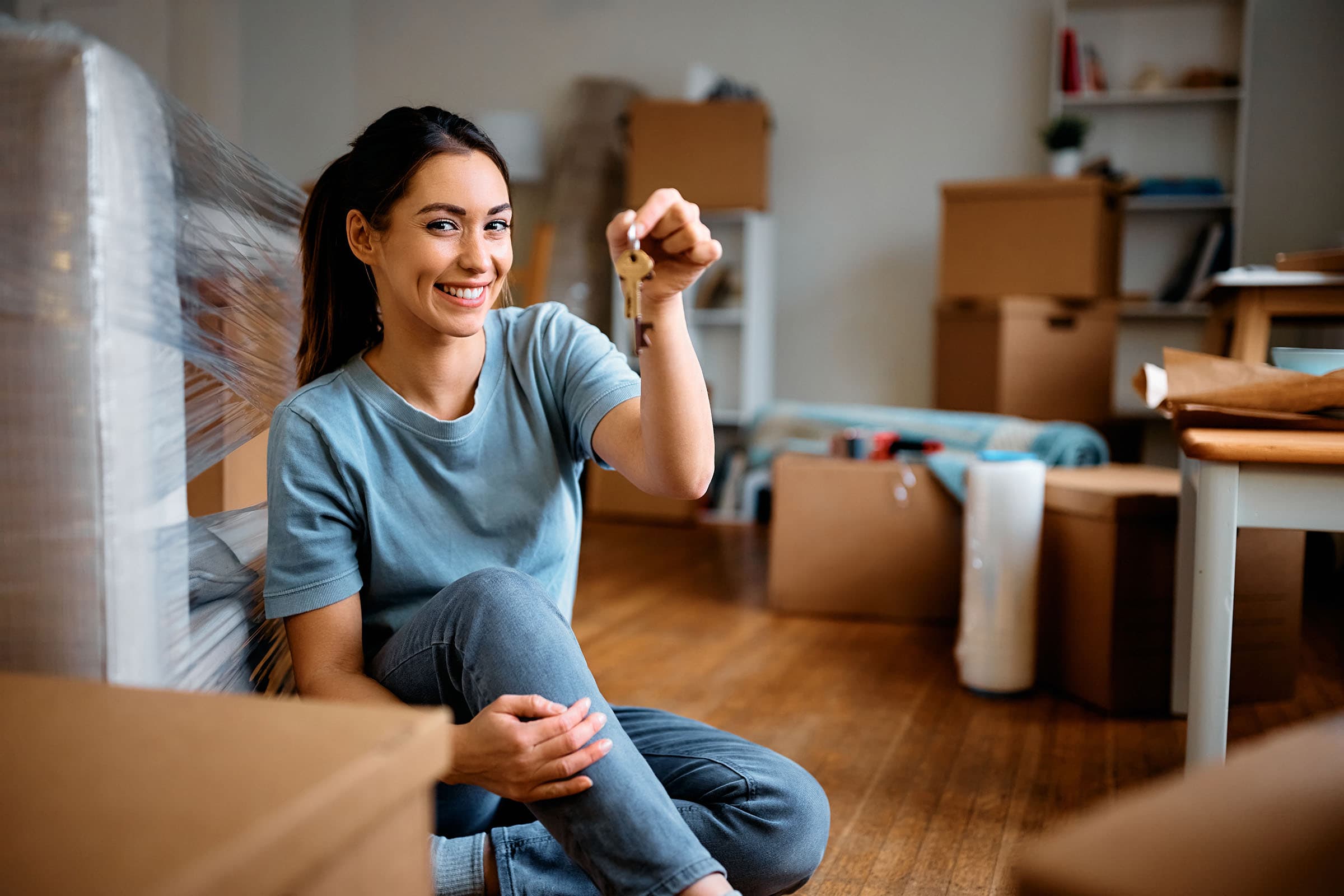 Smiling woman sitting on the floor holding house keys, surrounded by moving boxes and wrapped furniture in a new home.