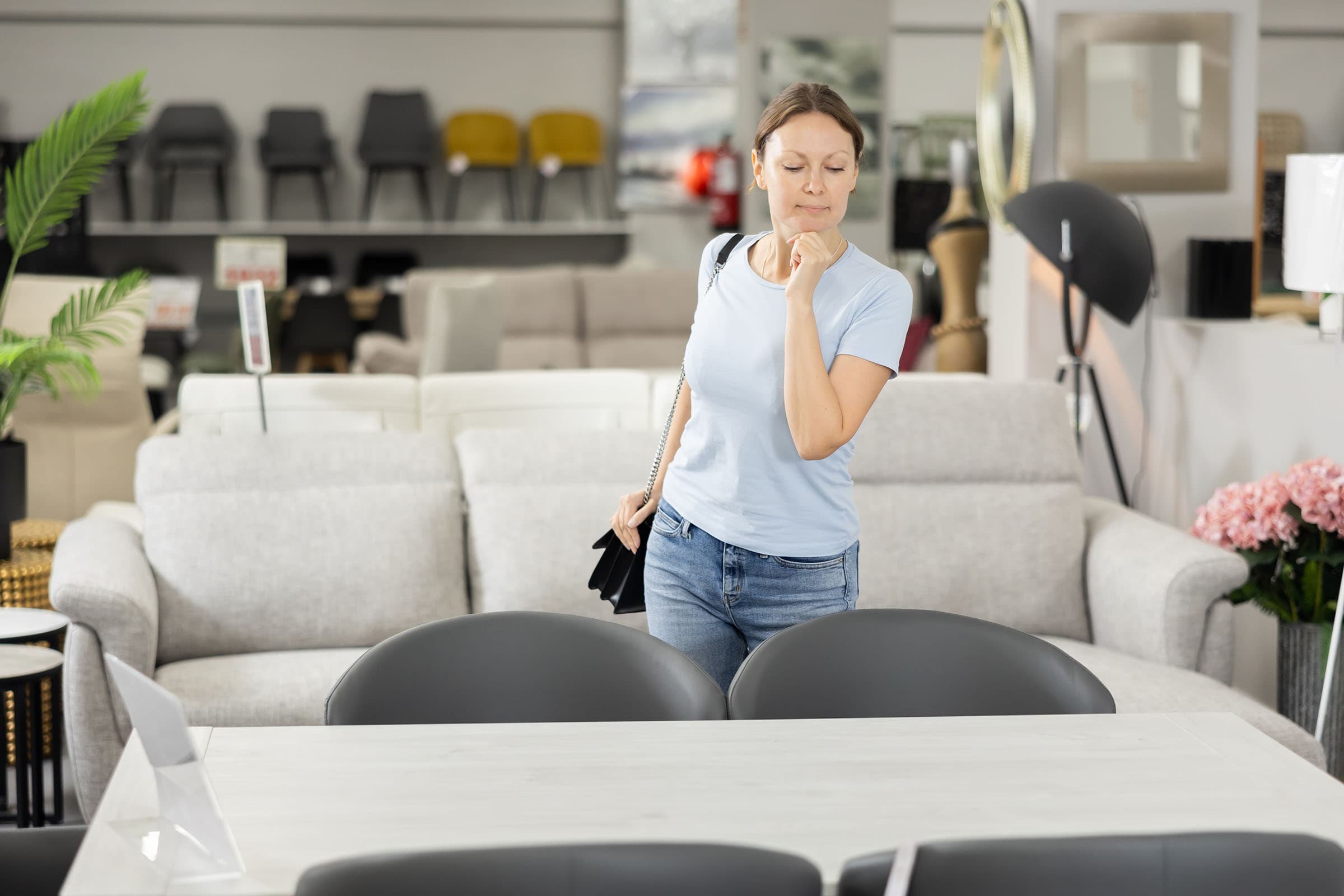 A woman in a furniture store is thoughtfully examining a gray dining table with chairs. Sofas and decorative items are displayed in the background.