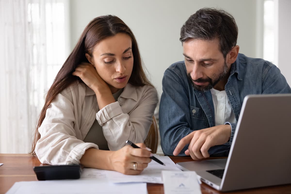 A man and woman sit at a table, discussing documents. The woman holds a pen while the man points at the papers. A laptop and calculator are nearby.
