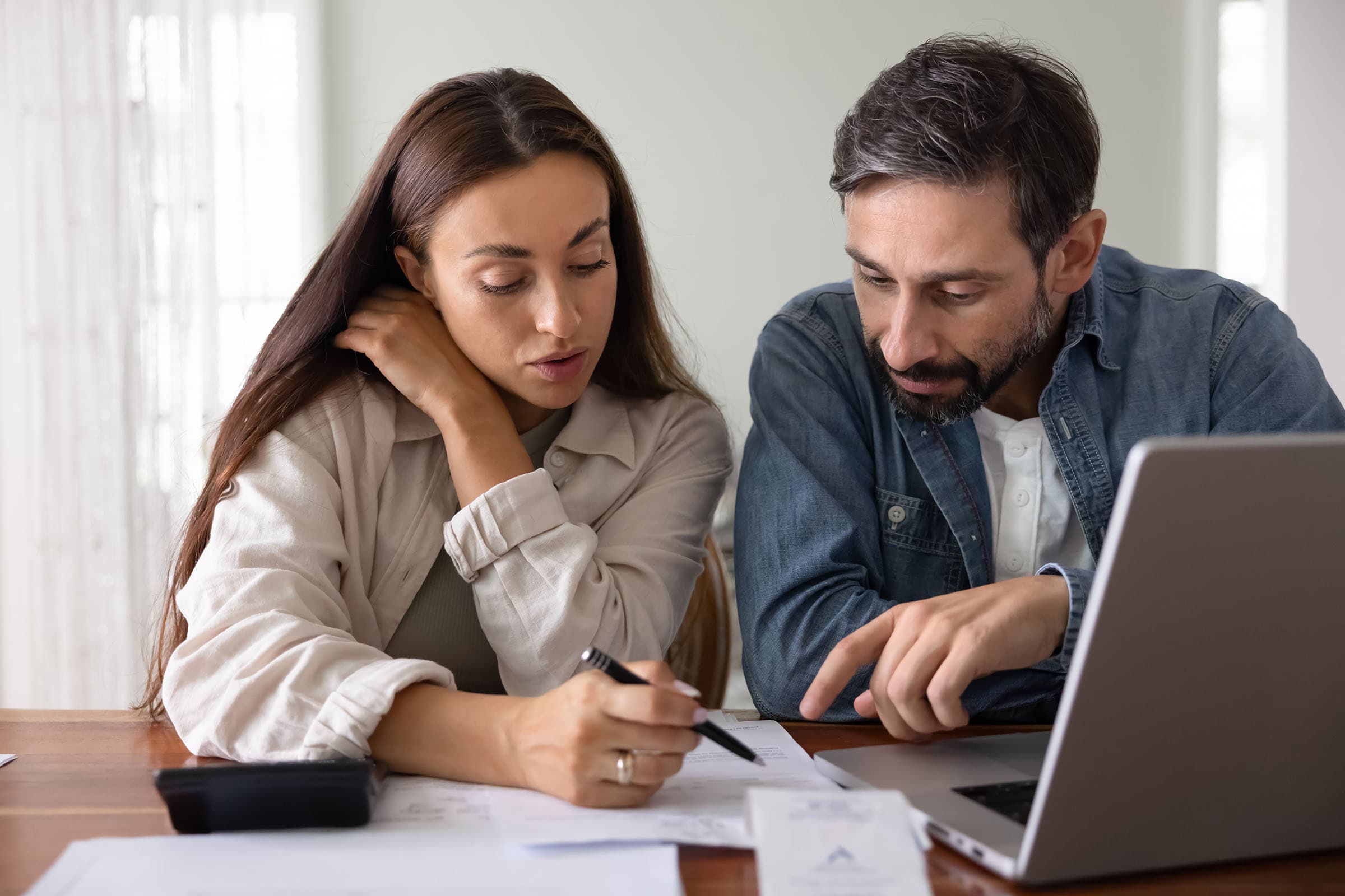 A man and woman sit at a table, discussing documents. The woman holds a pen while the man points at the papers. A laptop and calculator are nearby.
