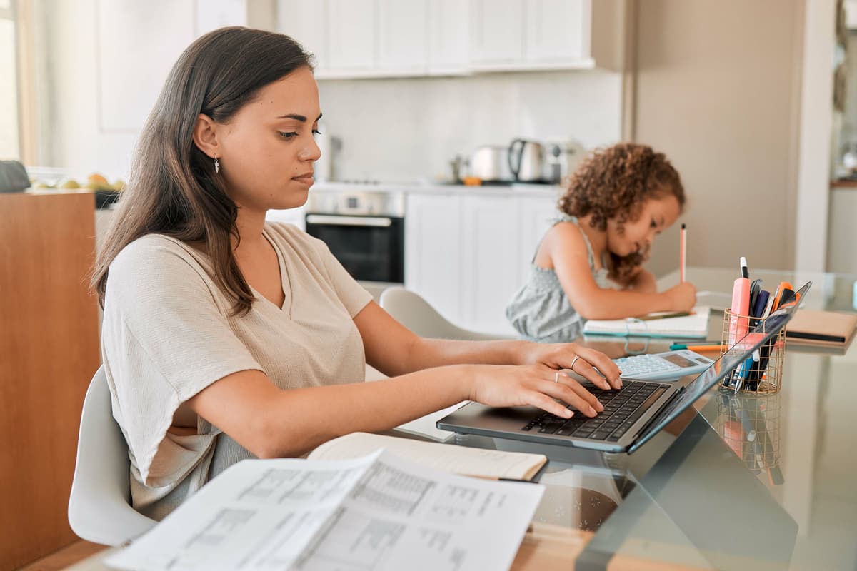 Woman working on a laptop at a kitchen table with papers and a calculator, while a young girl writes in a notebook beside her.