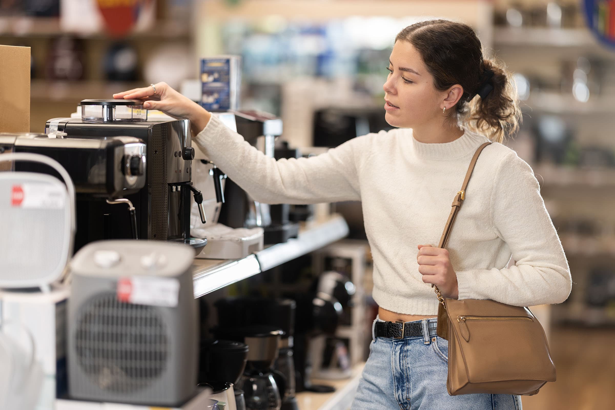 A woman in a white sweater and jeans examines a coffee machine in a store aisle, holding a brown shoulder bag.