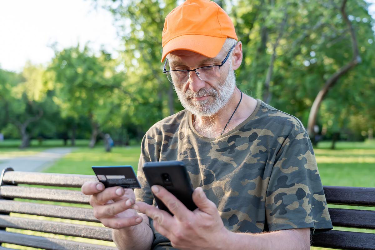 Older man in a camouflage shirt and orange cap sitting on a park bench, holding a credit card and using a smartphone.