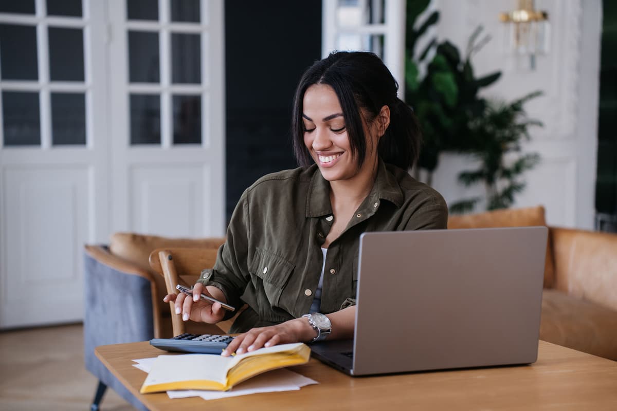 Smiling woman in a green shirt uses a calculator and notebook at a desk with a laptop. Cozy home setting with a sofa and plants in the background.