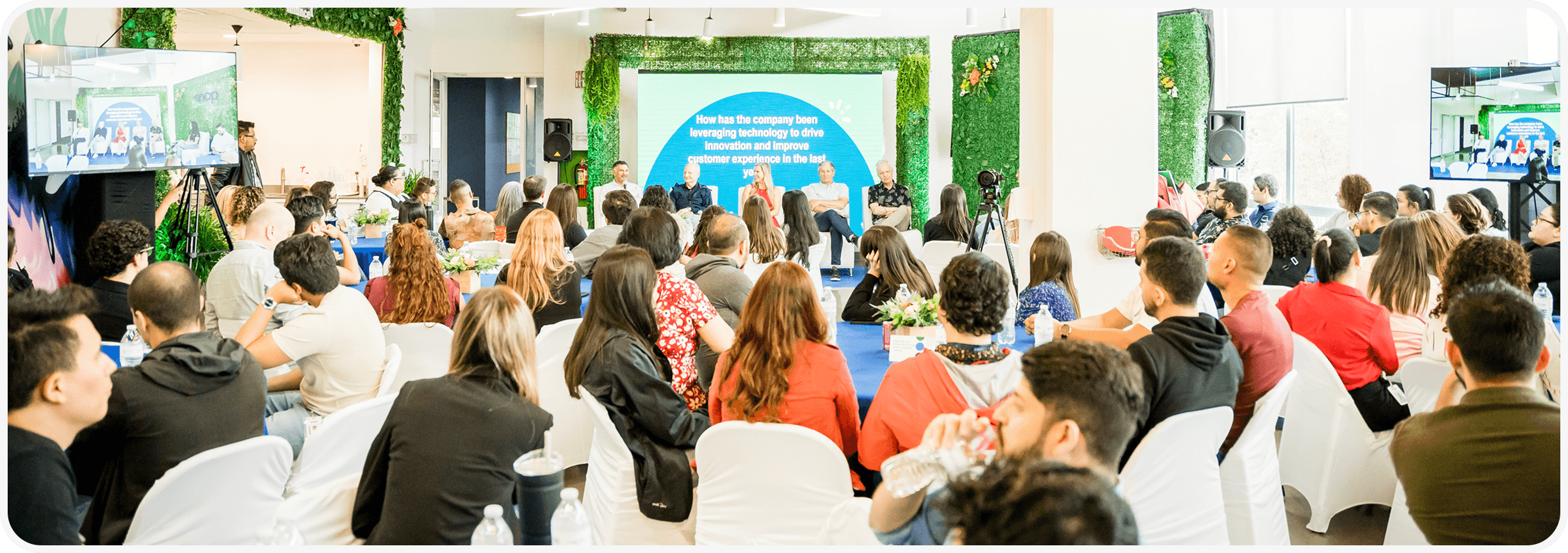 A diverse audience seated in a brightly lit room listens to a panel discussion. Screens display presentations, and greenery decorates the space.