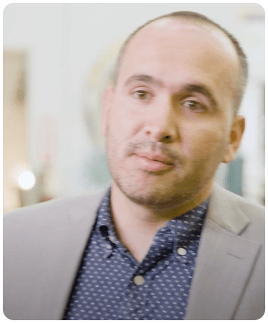A person with short hair and a beard, wearing a gray blazer over a patterned shirt, standing indoors with a blurred background.