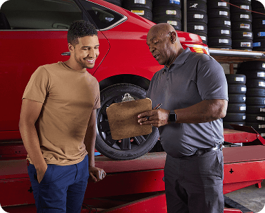 Two men in a garage, one holding a clipboard, standing near a red car on a lift, surrounded by stacked tires.