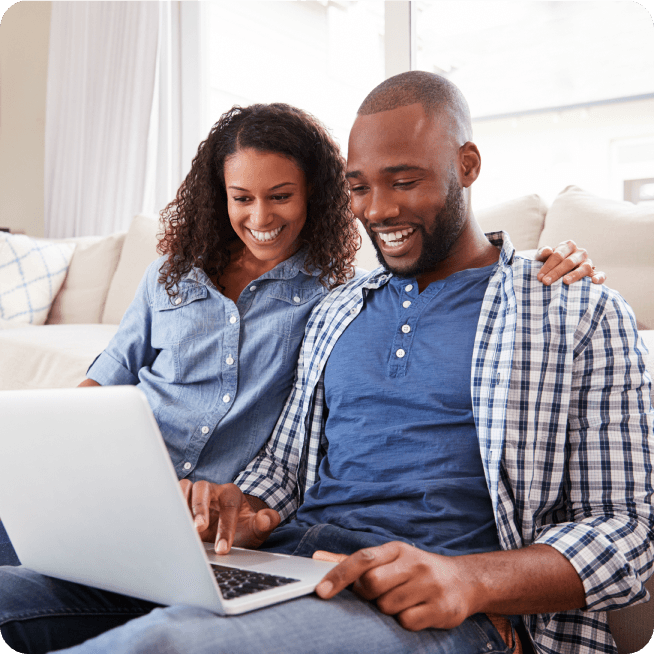 A smiling couple sits on a couch, looking at a laptop together, appearing happy and engaged in a bright, cozy living room.
