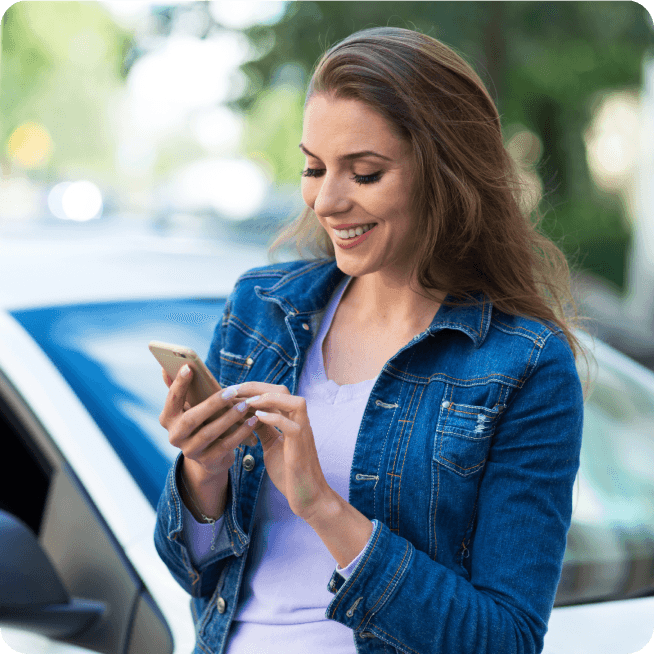 Smiling woman in a denim jacket using her smartphone outdoors near a car.