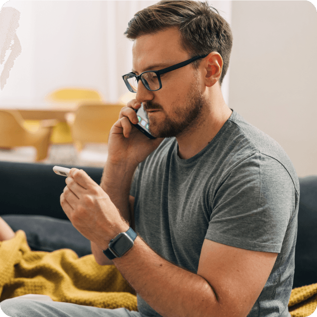 Man with glasses and beard, on the phone, holding and examining an object, sitting on a couch with a yellow blanket in a well-lit room.