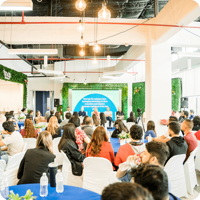 A group of people seated in a bright, modern conference room, attentively listening to a panel discussion in front of a large screen.