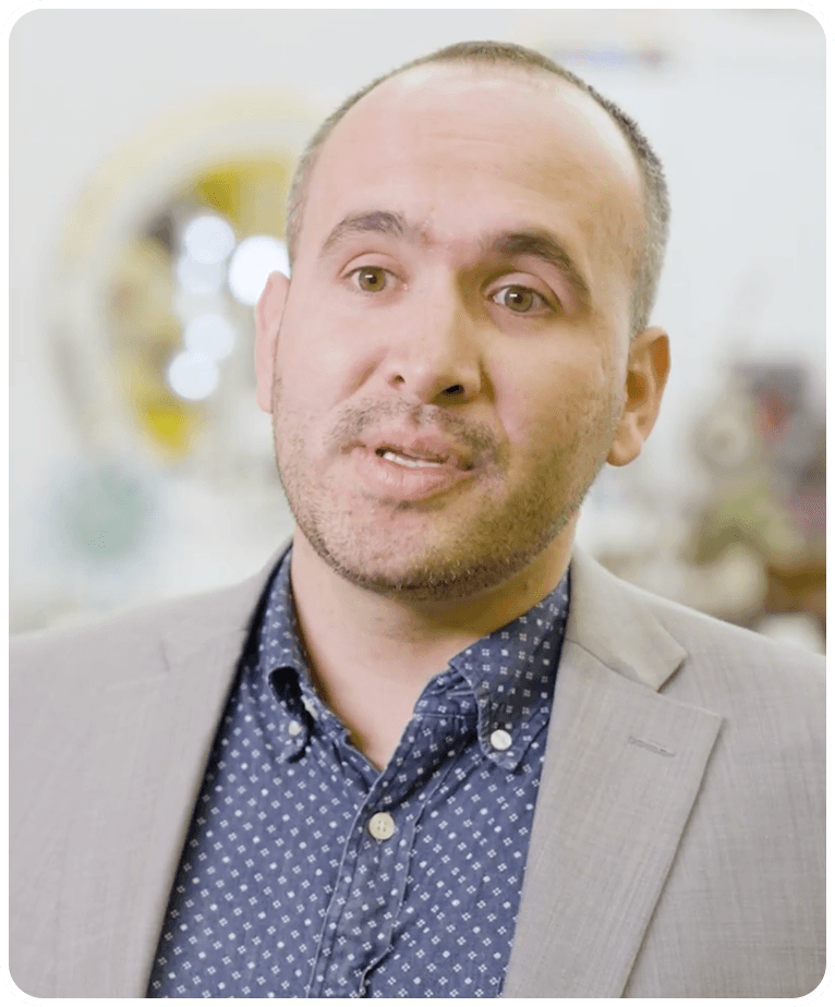 A man with a beard and short hair in a light gray suit and blue patterned shirt speaks, with a blurred background.