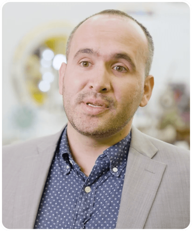 A man with a beard and short hair in a light gray suit and blue patterned shirt speaks, with a blurred background.