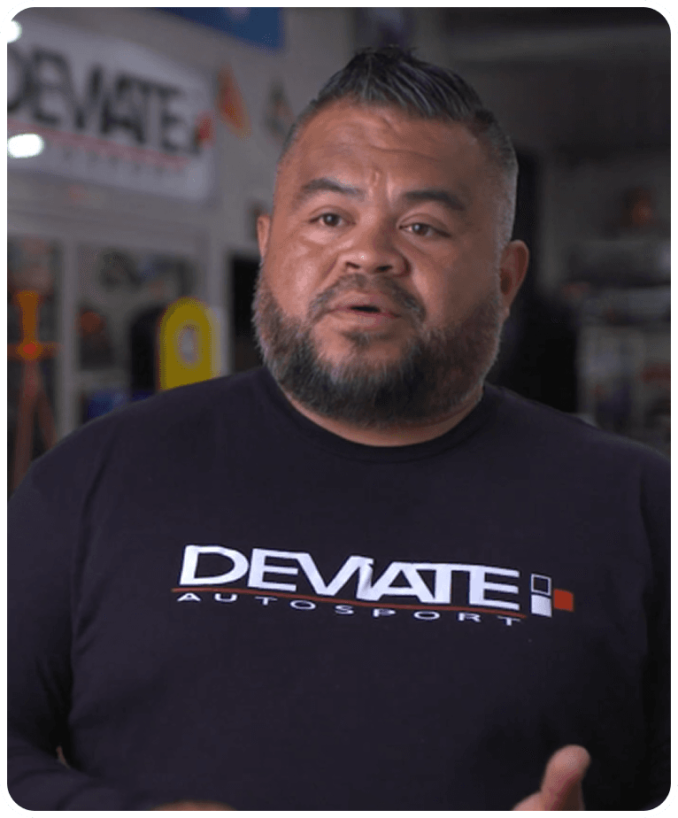 Man with a beard and short hair wearing a "DEVIATE AUTOSPORT" shirt, speaking in a workshop setting.