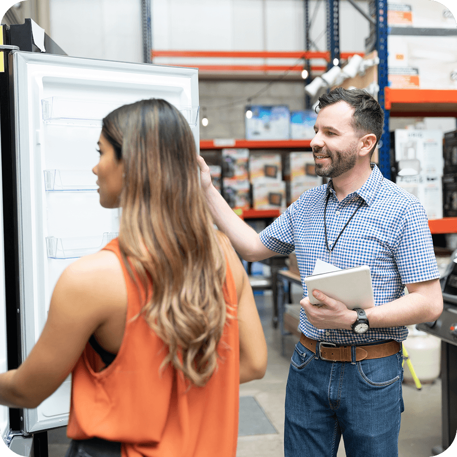 A salesperson shows a fridge to a customer in a store, holding a notepad. Shelves and product displays are in the background.