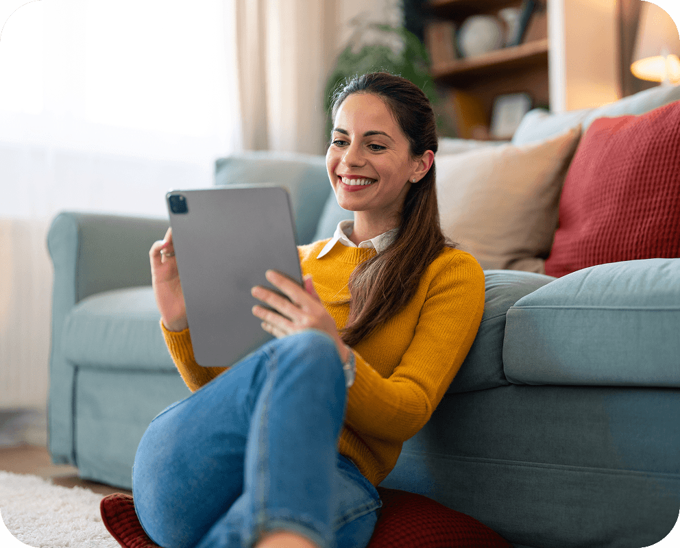 Smiling woman in a yellow sweater sitting on the floor, leaning against a sofa, using a tablet in a cozy living room.