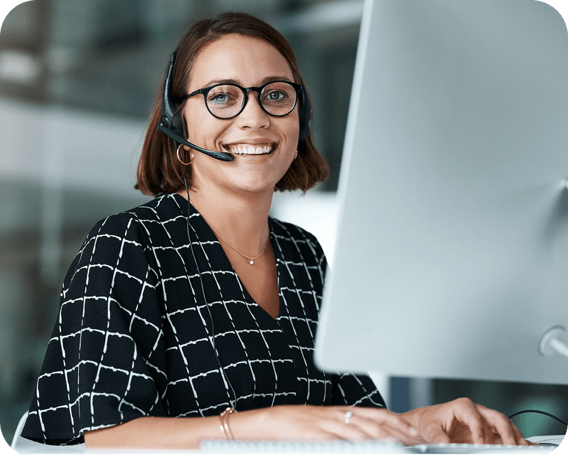 A woman with glasses and a headset smiles while working on a computer in an office environment.