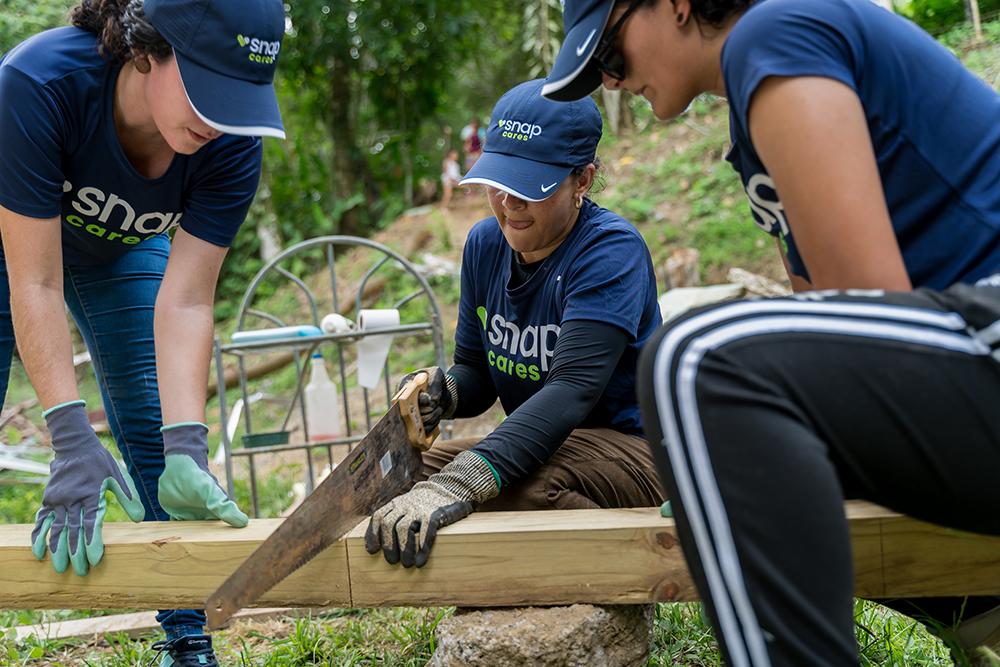 Three volunteers wearing blue 'Snap Cares' shirts work together on a community project in a lush, outdoor setting, using a saw to cut a wooden beam, showcasing teamwork and corporate social responsibility efforts.