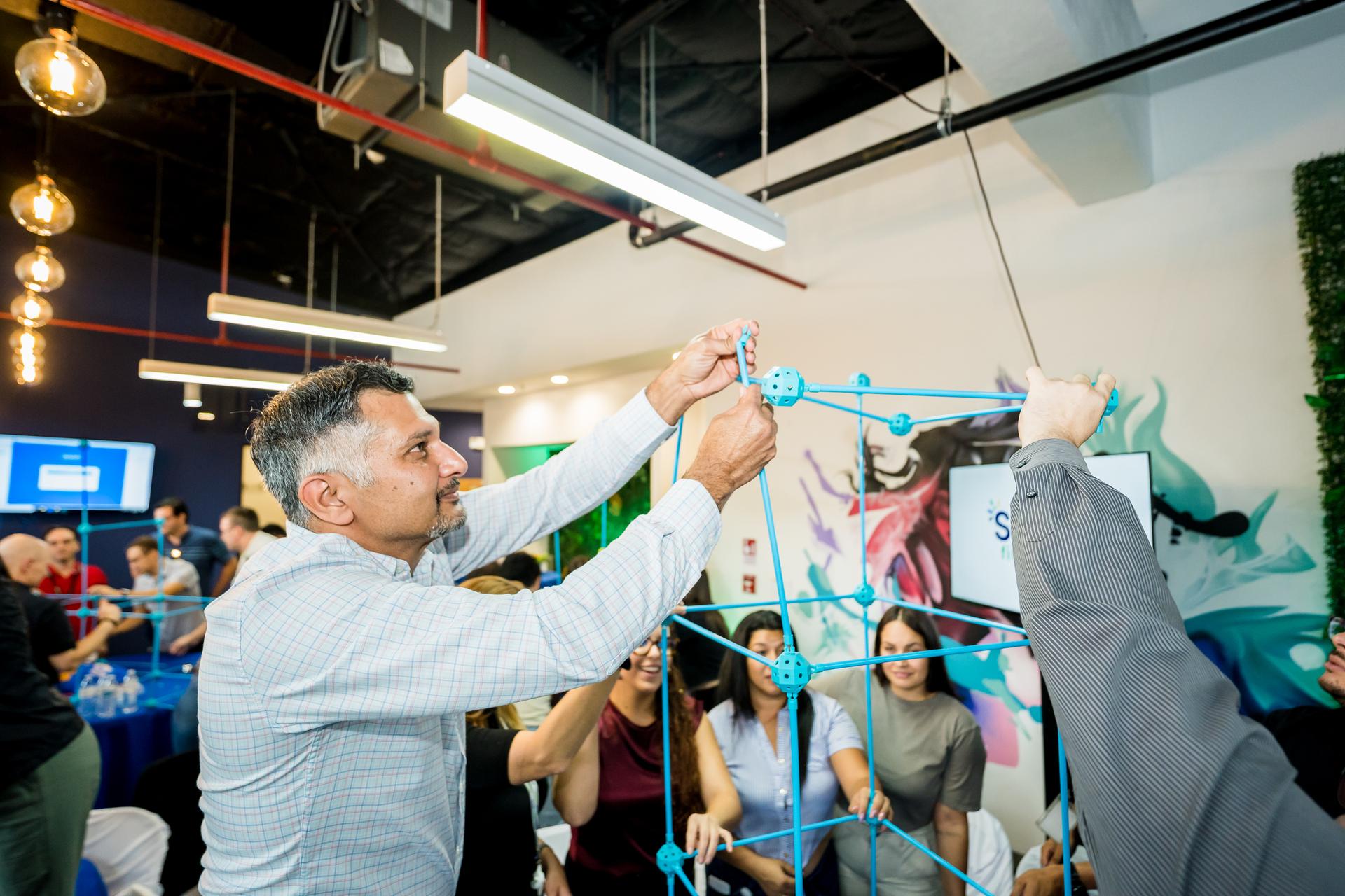 Employees collaborating in a colorful, modern office space, building a structure with blue plastic connectors during a team-building activity, highlighting creativity and teamwork in the workplace.