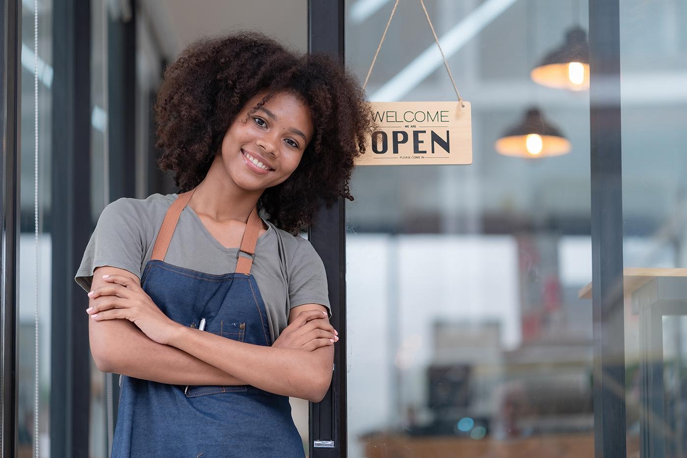 Small business owner smiling confidently at storefront, wearing denim apron and gray shirt, with 'Welcome Open' sign hanging in background