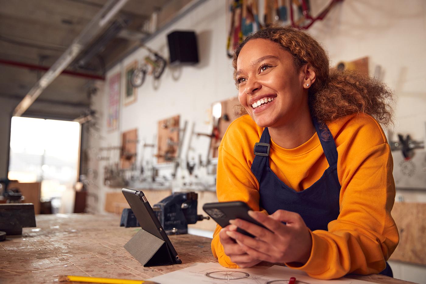 Young artisan or small business owner in bright yellow sweater and navy apron smiling while using tablet and phone in workshop, with tools and workspace visible in background