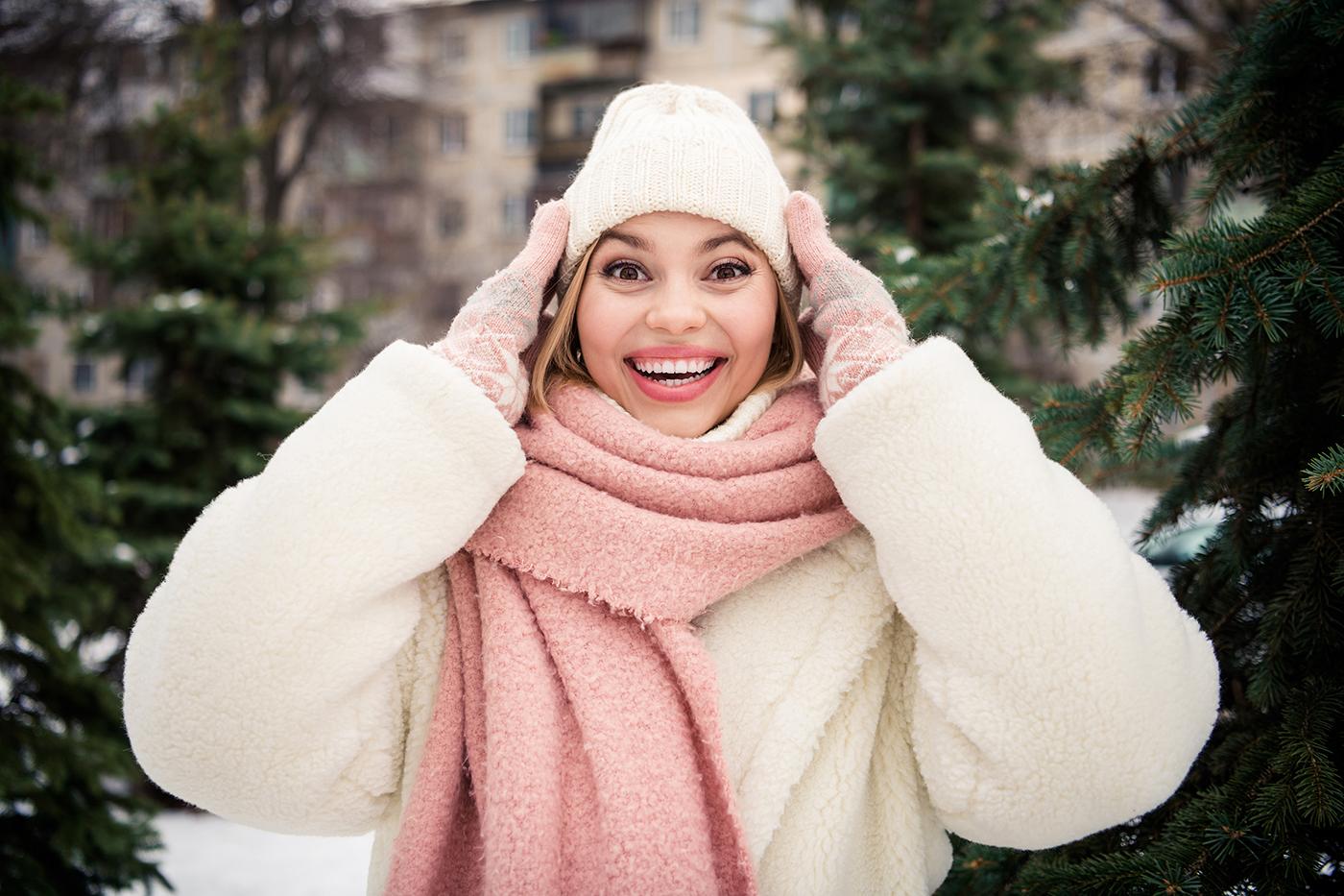 A woman in a winter coat and hat is smiling and laughing in a snowy outdoor setting with trees in the background.