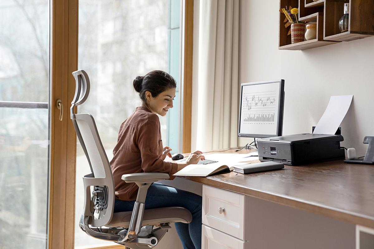Woman works at home-office desk on ergonomic chair; monitor shows data chart, printer and notebooks nearby.