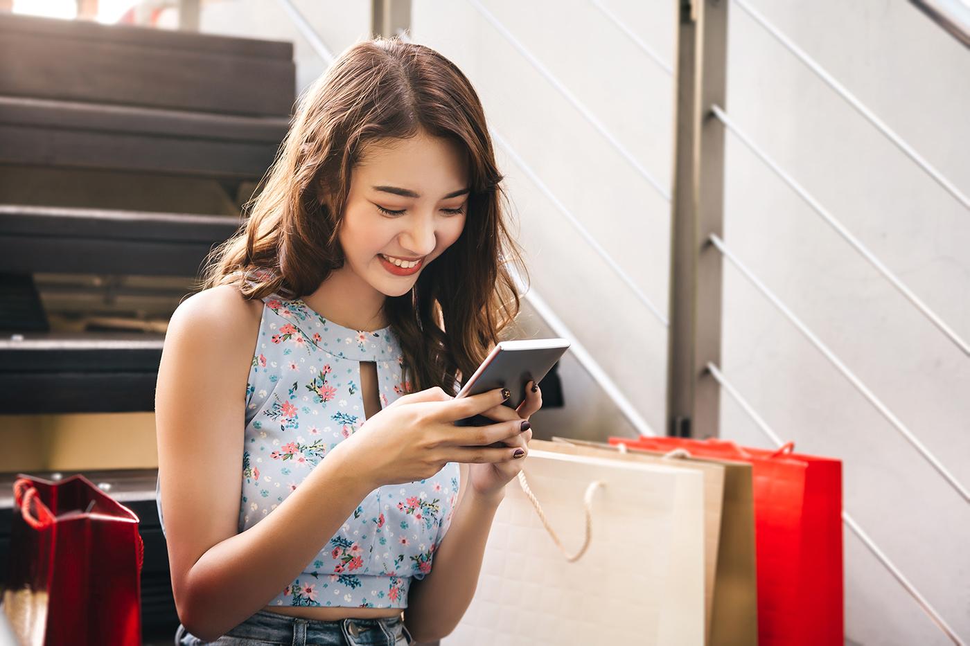 Young woman in floral sleeveless top sits on stairs, smiling at smartphone, surrounded by red and cream shopping bags.