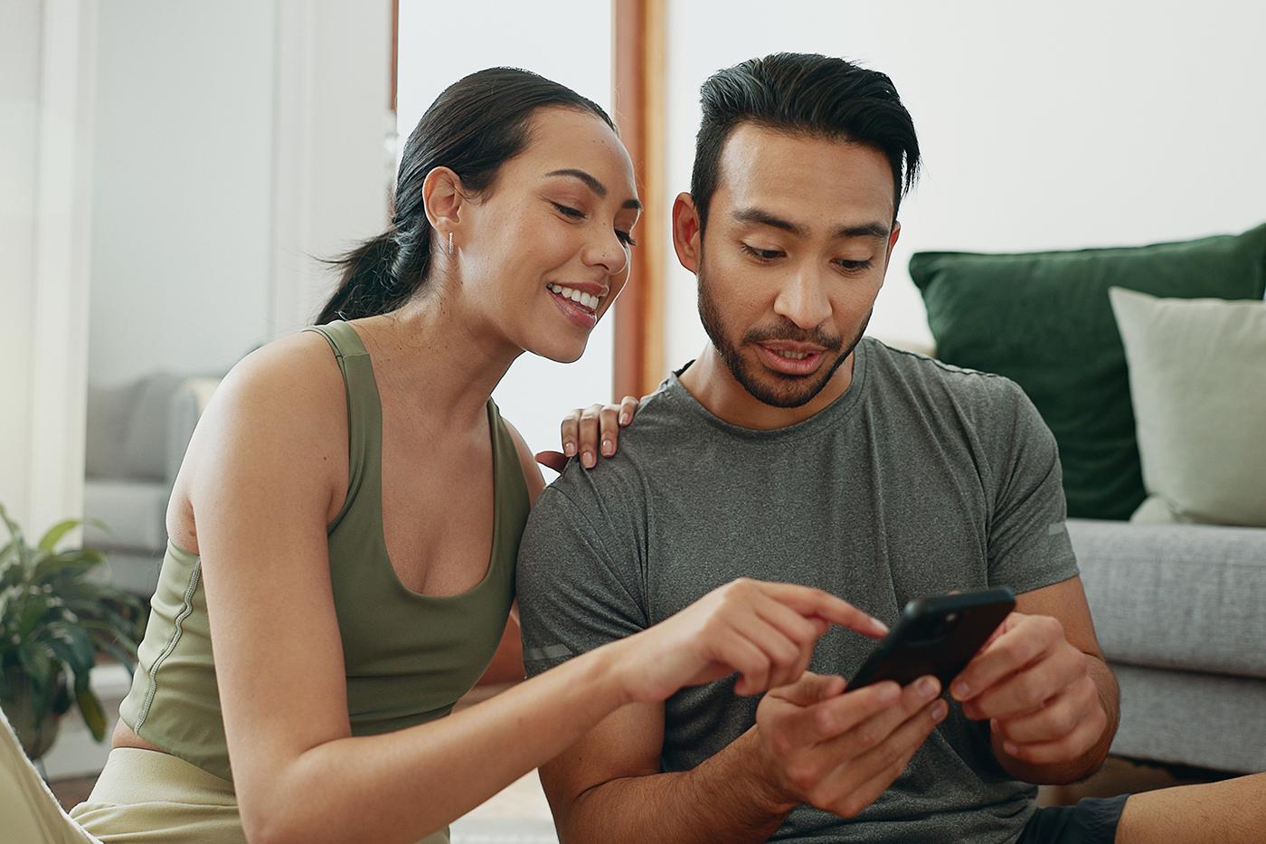 A woman and man sit indoors, smiling and looking at a smartphone the man is holding while the woman points at the screen.