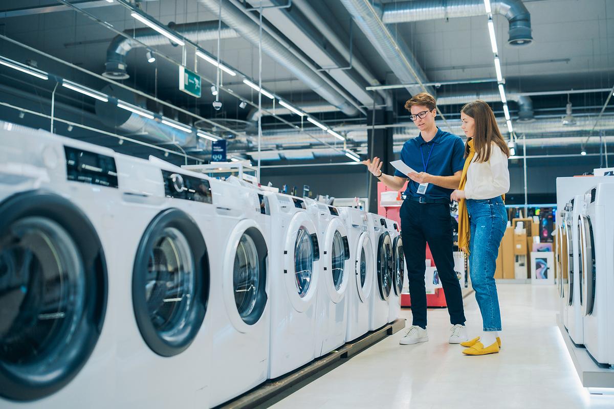 Store employee gestures while explaining a row of front-load washing machines to a woman shopper in a brightly lit appliance aisle.