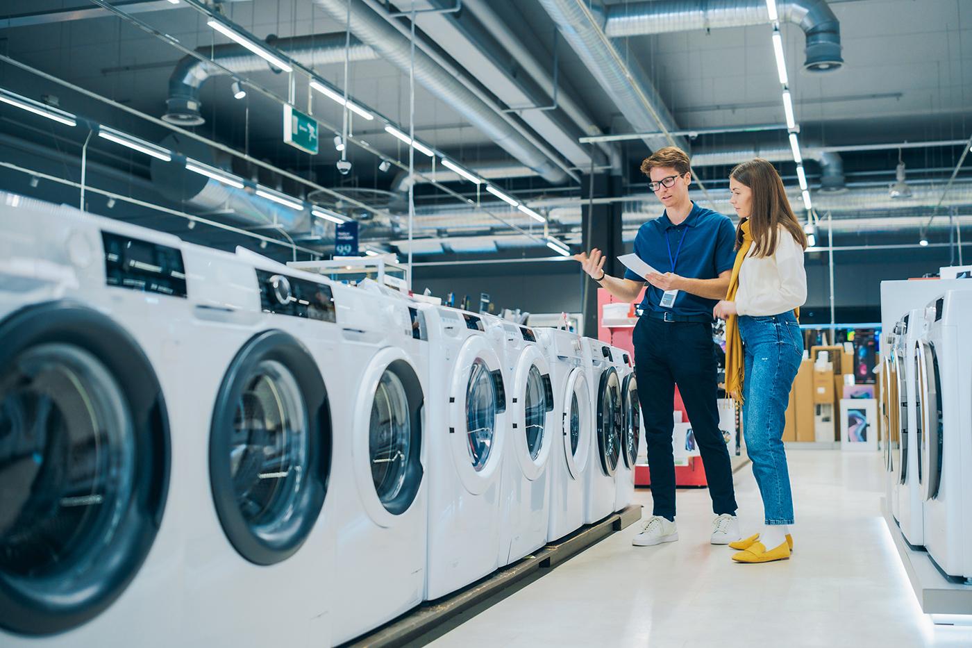 Store employee gestures while explaining a row of front-load washing machines to a woman shopper in a brightly lit appliance aisle.