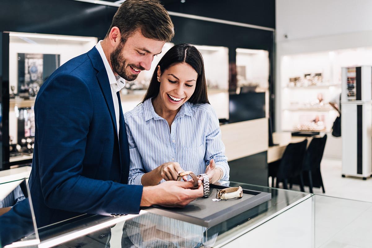 Smiling couple at glass display counter examine two wristwatches on a tray in a modern, well-lit jewelry store.