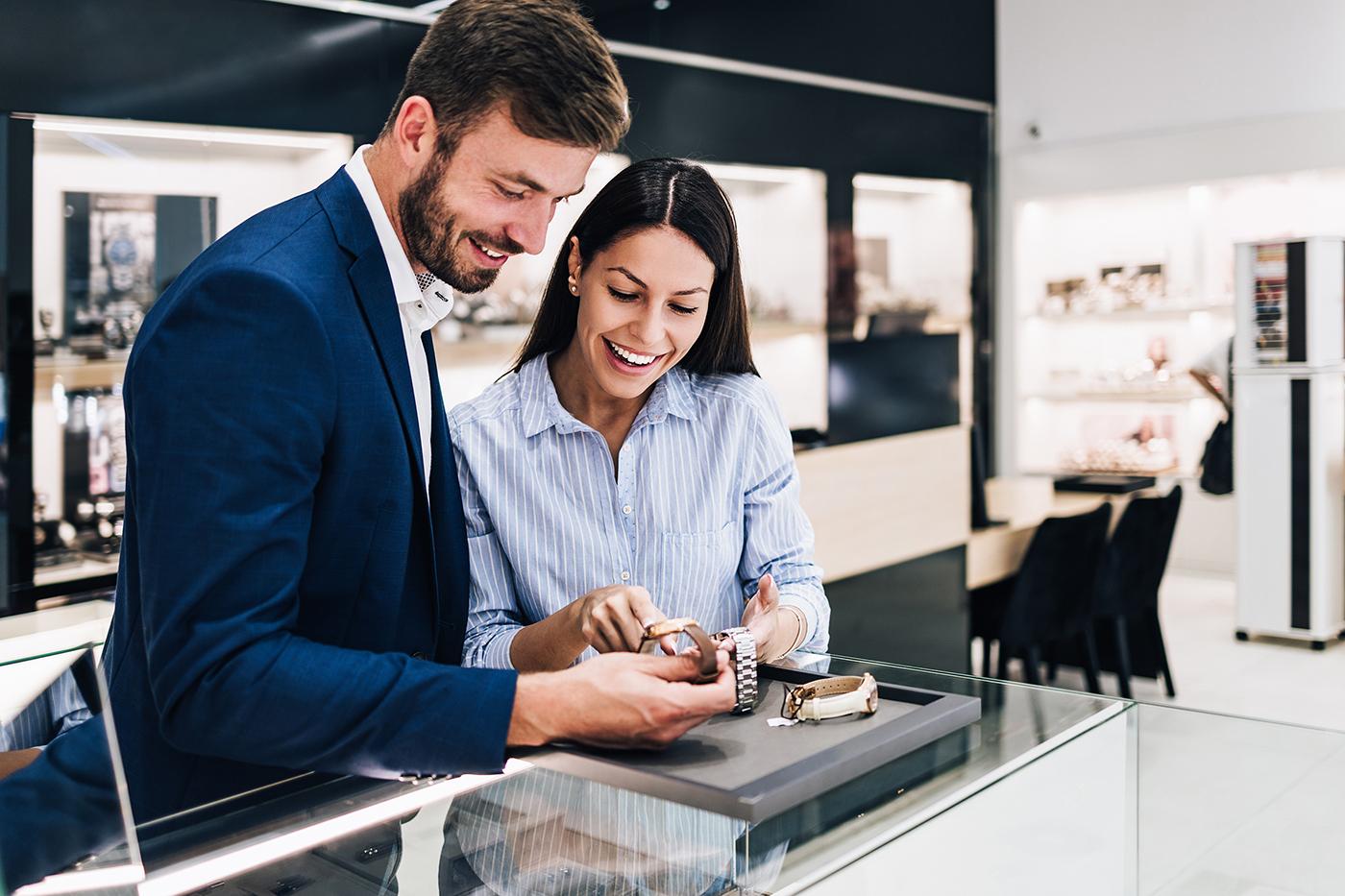 Smiling couple at glass display counter examine two wristwatches on a tray in a modern, well-lit jewelry store.