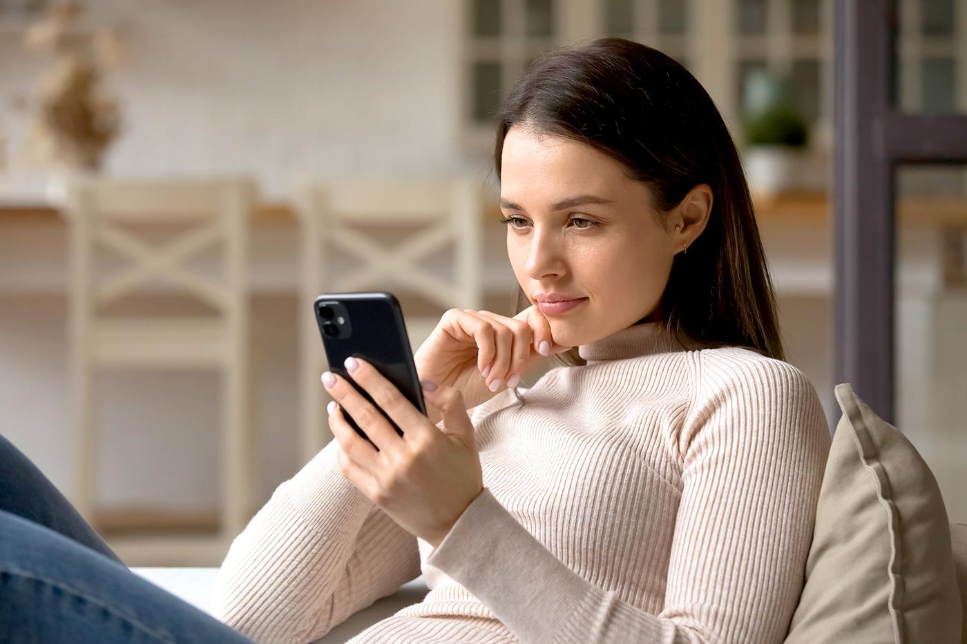 A woman sits on a couch, resting her chin on one hand while looking thoughtfully at a smartphone held in the other hand.