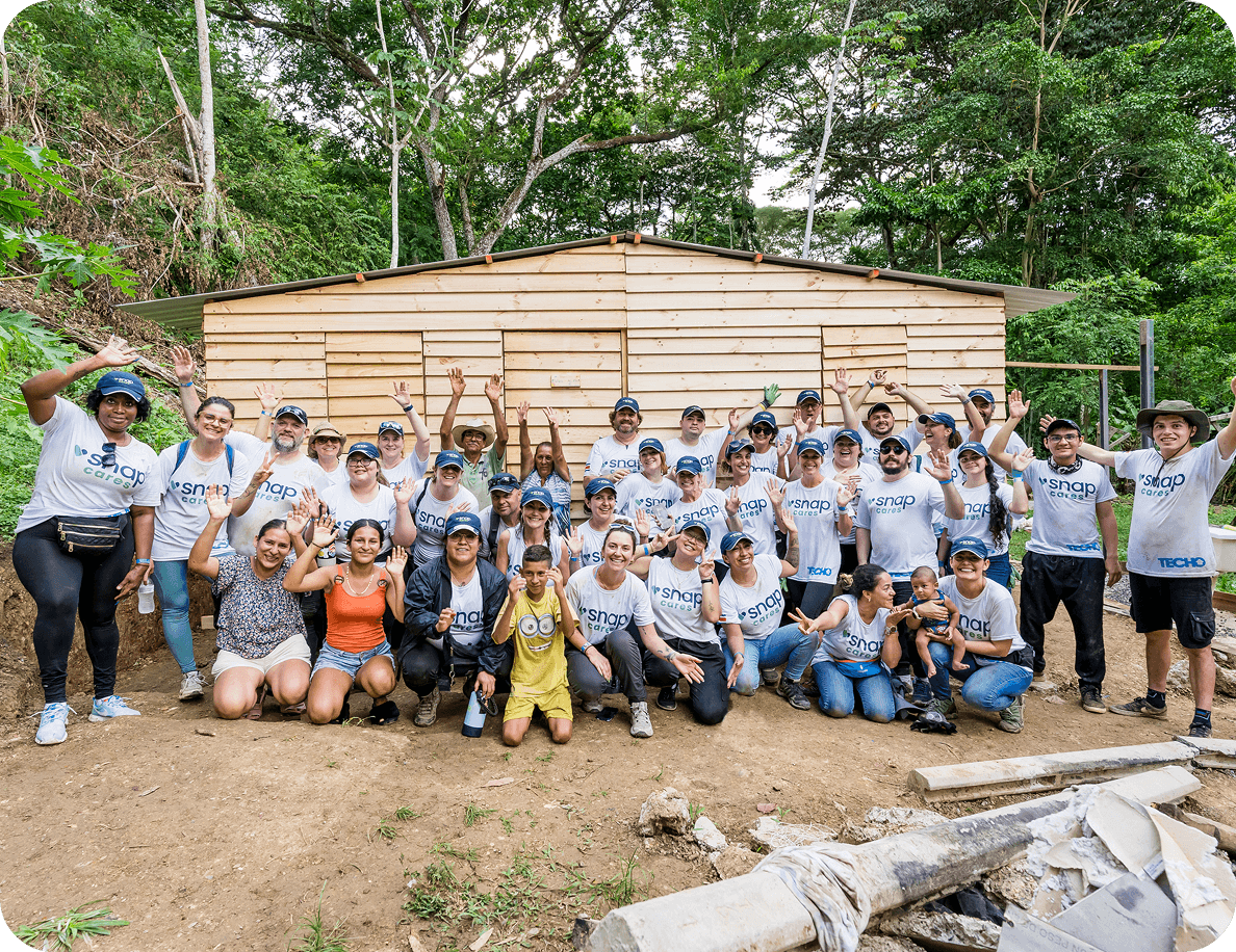 A large group of smiling people in matching shirts poses in front of a newly constructed wooden building, surrounded by lush greenery.