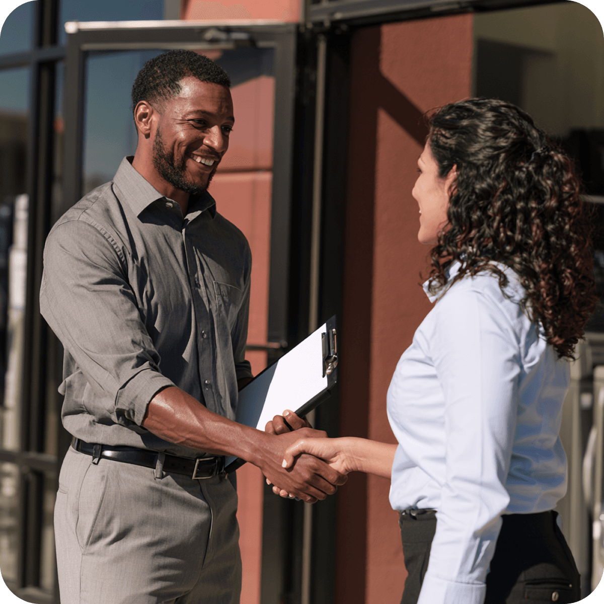Two people in business attire shaking hands outside a building, the man holding a clipboard.