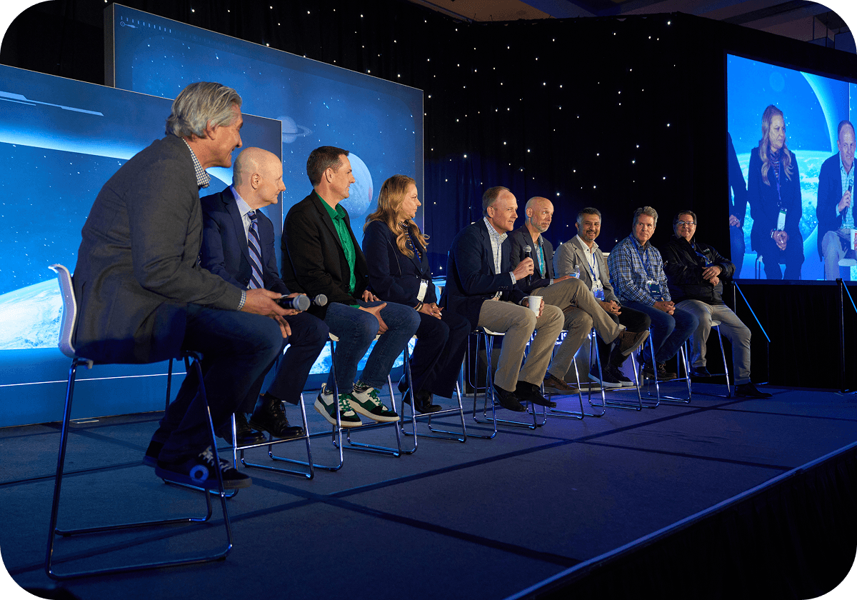 Nine people seated on a stage panel discussion, with a starry backdrop and a large screen displaying two of the panelists.